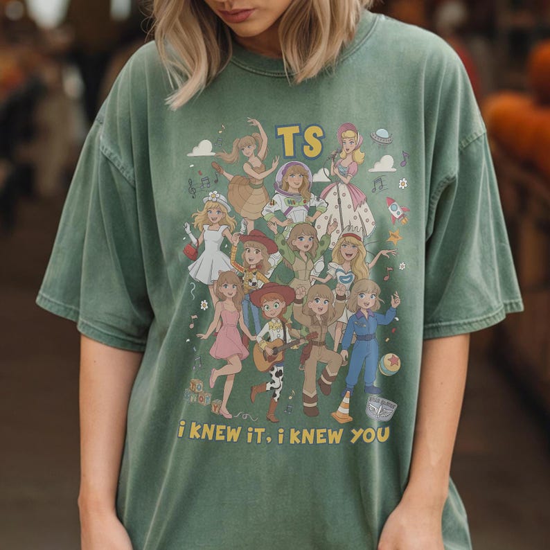 May include: A sage green short-sleeve t-shirt featuring a graphic print of multiple animated characters in various outfits. The text "TS" appears at the top, and the text "I KNEW IT, I KNEW YOU" is printed at the bottom in yellow lettering.