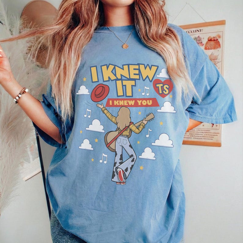 K&ouml;nnte beinhalten: Ein blaues Kurzarm-T-Shirt mit einer Grafik einer Person mit Gitarre, Wolken, Sternen und Musiknoten. Der Text lautet I KNEW IT I KNEW YOU TS in einem roten Herz. Die Person tr&auml;gt ein gelbes Oberteil und eine Hose mit Kuhfleckenmuster.