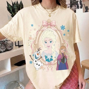 May include: Cream-colored t-shirt featuring a framed illustration of Elsa and Anna from Frozen, with Olaf the snowman. The design includes pink bows, snowflakes, and floral accents. The shirt is a casual wear item.
