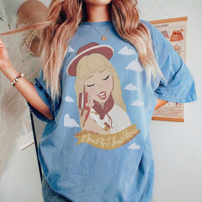 May include: A light blue t-shirt featuring a graphic illustration of a blonde person wearing a maroon hat and holding a small doll. The design includes white clouds and a banner with the cursive text I knew it, I knew you, printed in brown.