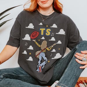 May include: A charcoal gray short-sleeve t-shirt featuring a graphic of a person with long hair wearing a yellow plaid shirt and cow-print bell-bottom jeans, holding a guitar and a red hat. The design includes white clouds, yellow stars, musical notes, and the letters TS.