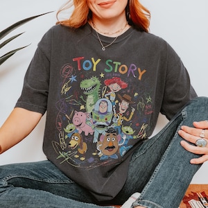 May include: A charcoal grey short-sleeve t-shirt featuring a colourful graphic print of Toy Story characters including Woody, Buzz Lightyear, Rex, Jessie, Slinky Dog, Mr. Potato Head, and Aliens, surrounded by multi-coloured scribbles and text.