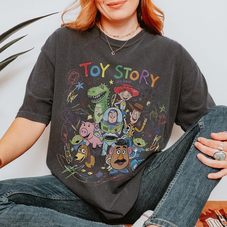 May include: A charcoal grey short-sleeve t-shirt featuring a colourful graphic print of Toy Story characters including Woody, Buzz Lightyear, Rex, Jessie, Slinky Dog, Mr. Potato Head, and Aliens, surrounded by multi-coloured scribbles and text.