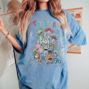 May include: A light blue t-shirt featuring a colourful graphic of Toy Story characters including Woody, Buzz Lightyear, Jessie, Rex, Hamm, Mr. Potato Head, Slinky Dog, and the Aliens, surrounded by scribbled star and line art designs.