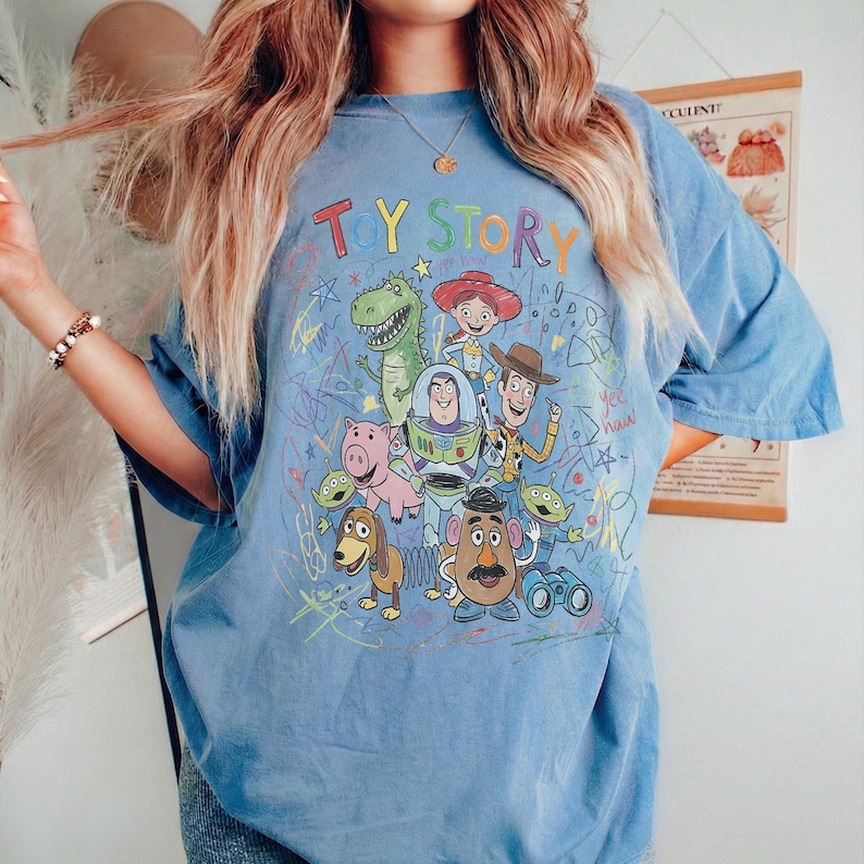 May include: A light blue t-shirt featuring a colourful graphic of Toy Story characters including Woody, Buzz Lightyear, Jessie, Rex, Hamm, Mr. Potato Head, Slinky Dog, and the Aliens, surrounded by scribbled star and line art designs.