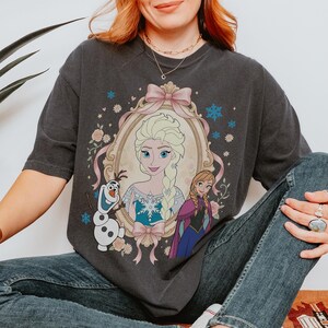 May include: Dark gray t-shirt featuring a framed portrait of Elsa from Frozen, with pink ribbon accents and floral details. The design includes images of Anna and Olaf. The shirt is a comfortable, casual style.