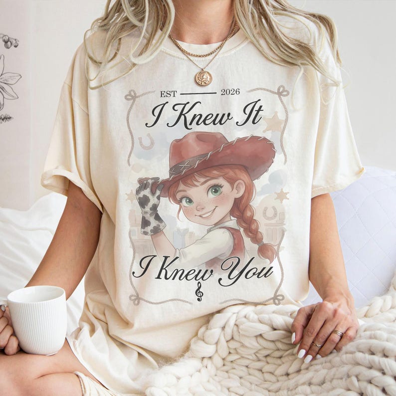 May include: A cream-colored t-shirt featuring a graphic of a cartoon cowgirl wearing a brown cowboy hat and a cow-print glove. The text reads "EST 2026", "I Knew It", and "I Knew You" with a small musical note symbol centered at the bottom.