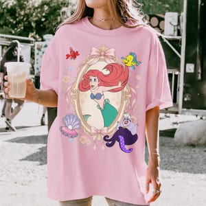 Puede incluir: Una camiseta rosa claro con una ilustraci&oacute;n de dibujos animados de Ariel de La Sirenita. El dise&ntilde;o incluye a Ariel en un espejo con marco dorado, junto con Flounder, Sebasti&aacute;n, &Uacute;rsula y otros elementos decorativos. La camiseta tiene mangas cortas.
