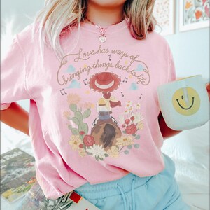 May include: A light pink t-shirt featuring a graphic of a cowgirl on a horse surrounded by cacti and flowers. The text above reads, Love has ways of bringing things back to life. The design includes musical notes, clouds, and stars in soft colors.