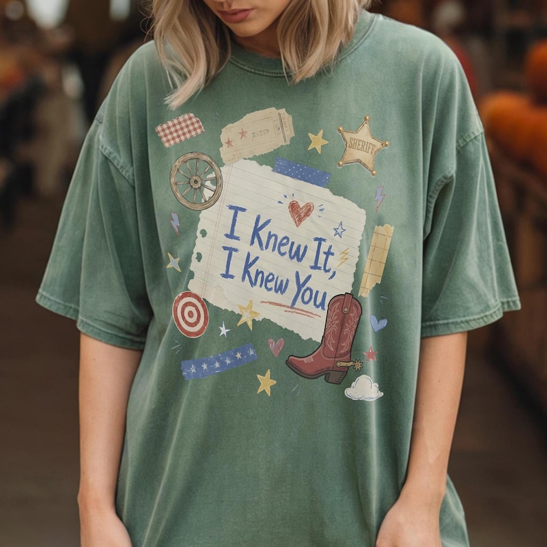 May include: A sage green short-sleeve t-shirt featuring a central graphic of a torn notebook page with the text "I Knew It, I Knew You" in blue. The design includes a brown cowboy boot, a sheriff star, a wagon wheel, and various small icons.