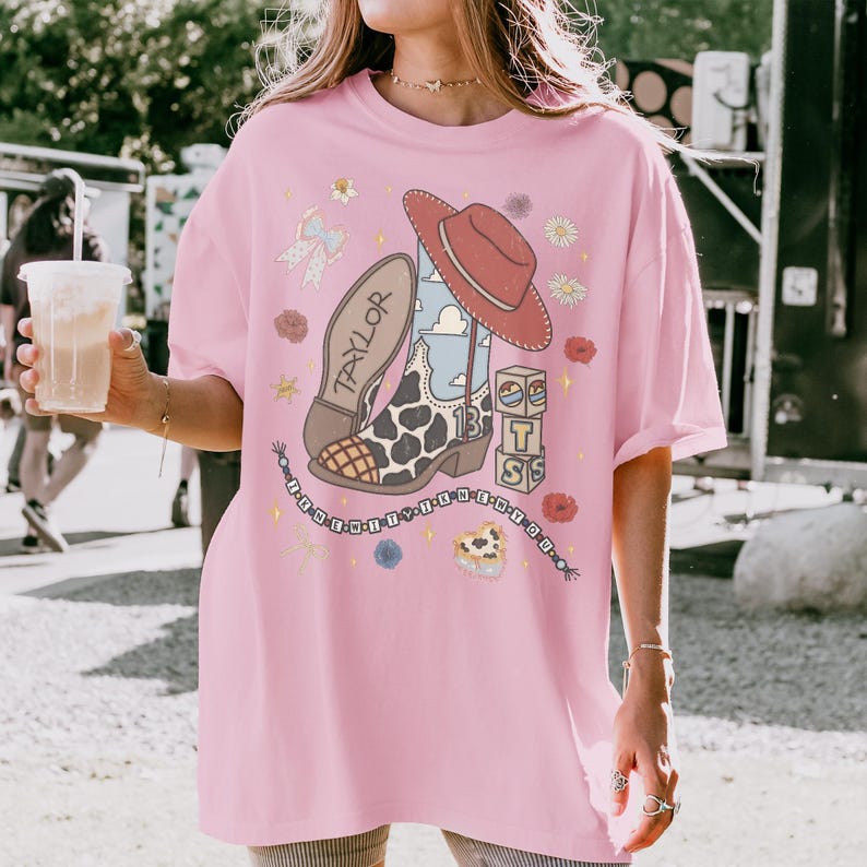 May include: A light pink short-sleeve t-shirt featuring a graphic print of a cowboy boot with a cow print pattern, a red hat, stacked blocks with the letters T S, and a beaded string with the text I KNEW YOU.