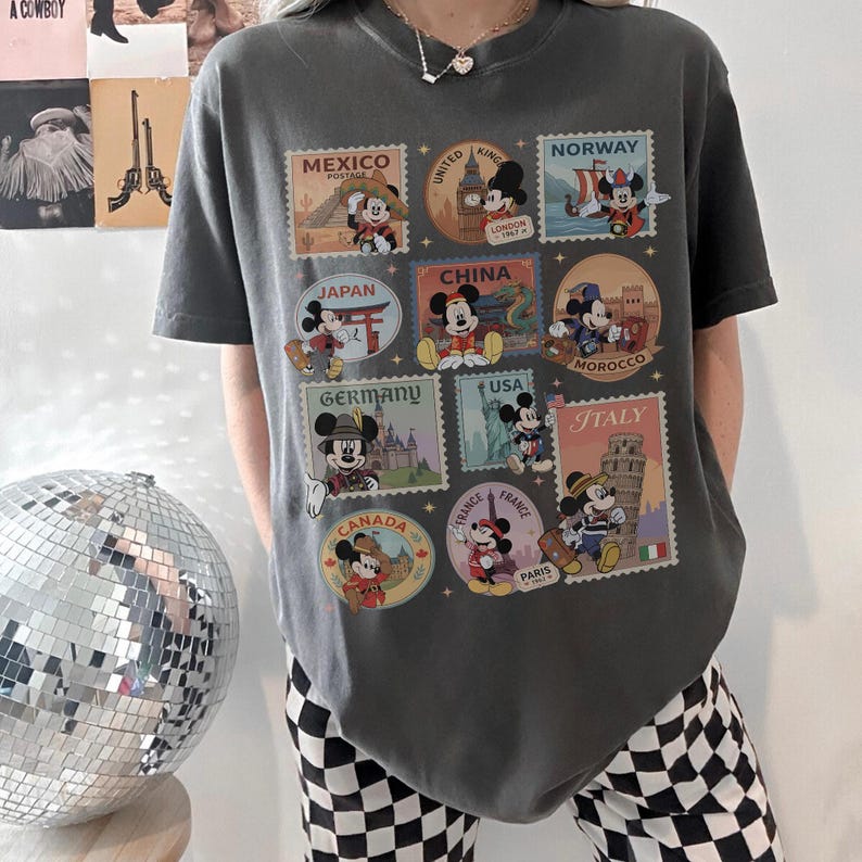 May include: Dark gray t-shirt featuring Mickey Mouse postage stamp designs from various countries, including Mexico, Japan, and Italy. The shirt has a crew neck and short sleeves. The shirt is a casual style.
