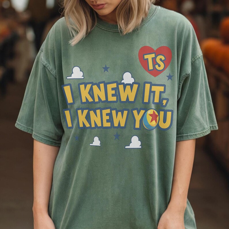 Op de afbeelding: Een saliegroen t-shirt met korte mouwen, voorzien van een rood hart met de letters TS, de tekst I KNEW IT, I KNEW YOU in het geel met blauwe omlijning, een kleine balafbeelding met een rode ster, en witte wolken en blauwe sterren.