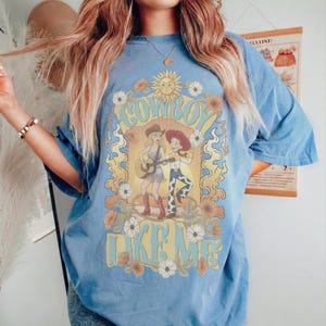 May include: A blue short-sleeve t-shirt featuring a graphic print of two female figures, one playing a guitar, surrounded by sunflowers and a sun. The text "COWBOY LIKE ME" is printed in a large, stylized yellow font above and below the illustration.