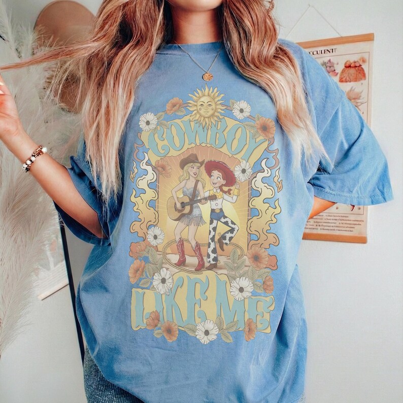 May include: A blue short-sleeve t-shirt featuring a graphic print of two female figures, one playing a guitar, surrounded by sunflowers and a sun. The text "COWBOY LIKE ME" is printed in a large, stylized yellow font above and below the illustration.
