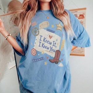 May include: A light blue short-sleeve t-shirt featuring a central graphic of a torn piece of paper with the text 'I Knew It, I Knew You'. The design includes a brown cowboy boot, a sheriff star, a wagon wheel, a target, and small stars.