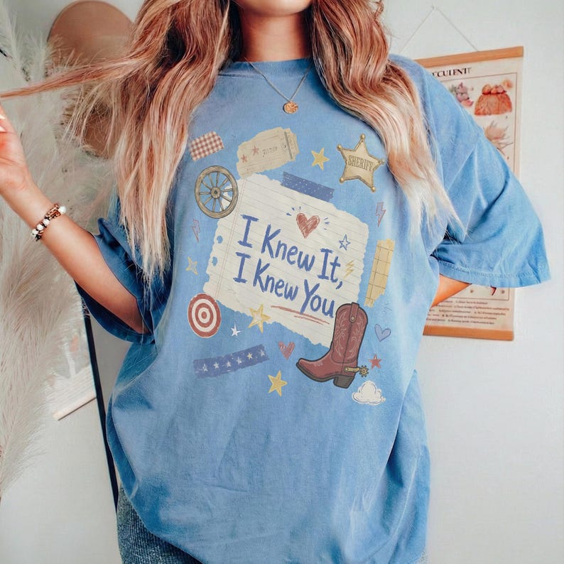 May include: A light blue short-sleeve t-shirt featuring a central graphic of a torn piece of paper with the text 'I Knew It, I Knew You'. The design includes a brown cowboy boot, a sheriff star, a wagon wheel, a target, and small stars.