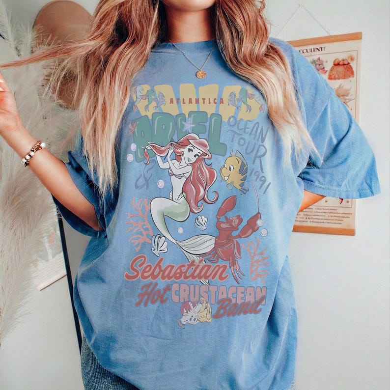 May include: Light blue t-shirt featuring a graphic of Ariel from The Little Mermaid, with the text "Atlantica Ocean Tour 1991." The design includes illustrations of Ariel, Flounder, and Sebastian. The shirt has short sleeves and a relaxed fit.