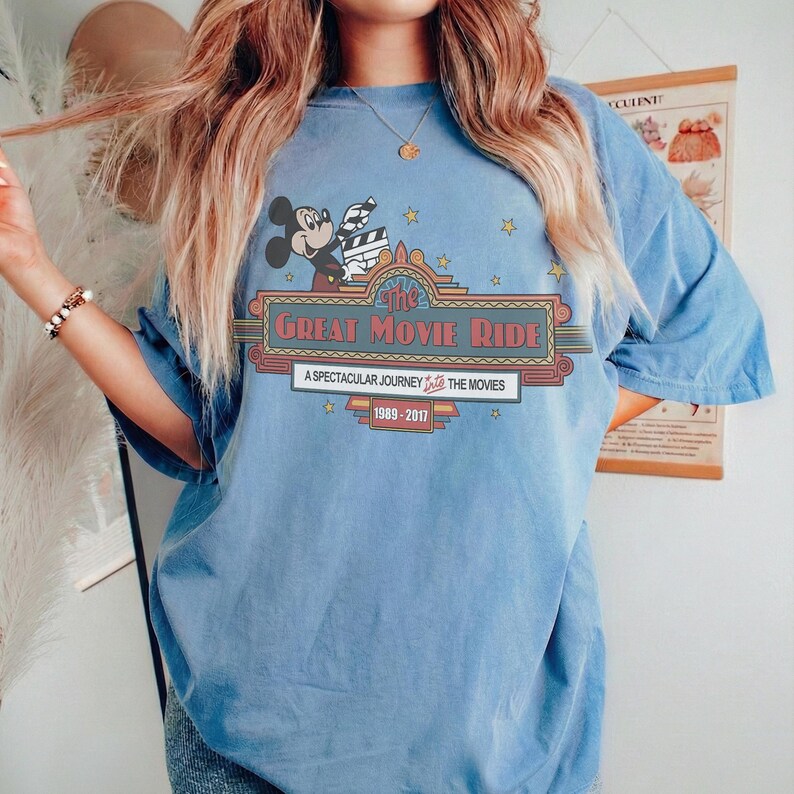 May include: A pale blue t-shirt with a graphic of Mickey Mouse holding a film clapperboard above the words "The Great Movie Ride". The design includes stars and the text "A Spectacular Journey Into The Movies 1989-2017".