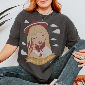 May include: A charcoal gray short-sleeve t-shirt featuring a graphic illustration of a blonde character wearing a red hat and holding a smaller doll. The design includes white clouds and a gold banner with the text I Knew It, I Knew You.