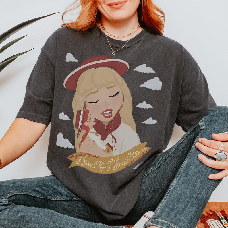 May include: A charcoal gray short-sleeve t-shirt featuring a graphic illustration of a blonde character wearing a red hat and holding a smaller doll. The design includes white clouds and a gold banner with the text I Knew It, I Knew You.
