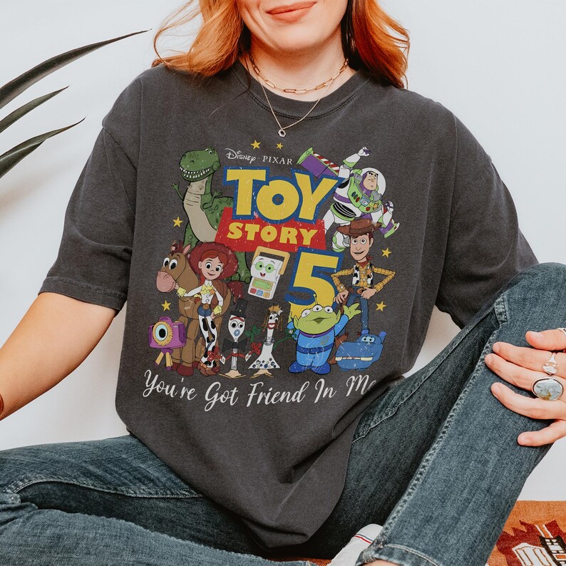 May include: A charcoal gray short-sleeve t-shirt featuring a colorful graphic print of Toy Story characters including Woody, Buzz Lightyear, Jessie, and Rex. The text reads Disney Pixar Toy Story 5 and You've Got Friend In Me with yellow stars.