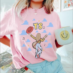 May include: A light pink t-shirt featuring a graphic of a person with long hair wearing a yellow plaid shirt and cow-print pants, holding a guitar and a red hat. The design includes blue clouds, musical notes, yellow stars, and the letters TS.