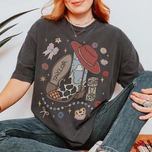 May include: A charcoal grey short-sleeve t-shirt featuring a graphic print of a cow-print cowboy boot, a red hat, a bow, flowers, and blocks with the letters T, S, and the number 13. The text "TAYLOR" is on the boot sole and "I KNEW IT" is below.