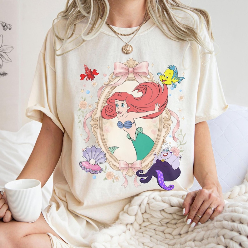 Puede incluir: Camiseta color crema con una ilustraci&oacute;n de dibujos animados de una sirena en un marco decorativo, rodeada de elementos con tem&aacute;tica marina. El dise&ntilde;o incluye una langosta roja, un pez amarillo, una concha y un pulpo morado. La sirena tiene pelo rojo y cola verde.