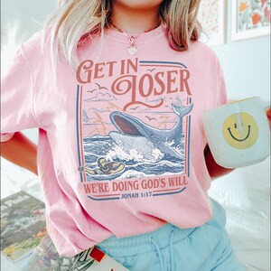 Puede incluir: Camiseta rosa claro con un gr&aacute;fico de una ballena rompiendo la superficie del oc&eacute;ano, con el texto "GET IN LOSER" y "WE'RE DOING GOD'S WILL" y el vers&iacute;culo b&iacute;blico Jon&aacute;s 1:17. Una taza blanca con una carita sonriente amarilla se sostiene en la mano.