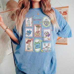 Puede incluir: Una camiseta azul de manga corta con un estampado gr&aacute;fico de varios personajes de Toy Story dispuestos en marcos estilo sello postal. El texto central dice You've Got A Friend In Me, rodeado de ilustraciones de Woody, Buzz Lightyear, Jessie, Forky y otros personajes.