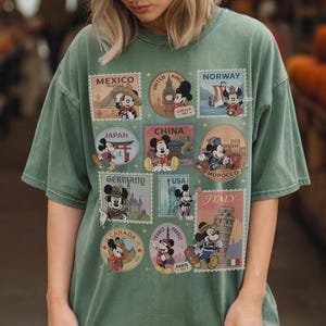May include: Sage green t-shirt featuring Mickey Mouse postage stamp designs representing various countries, including Mexico, Japan, China, Germany, USA, Italy, and France. The stamps showcase Mickey in different outfits and settings.