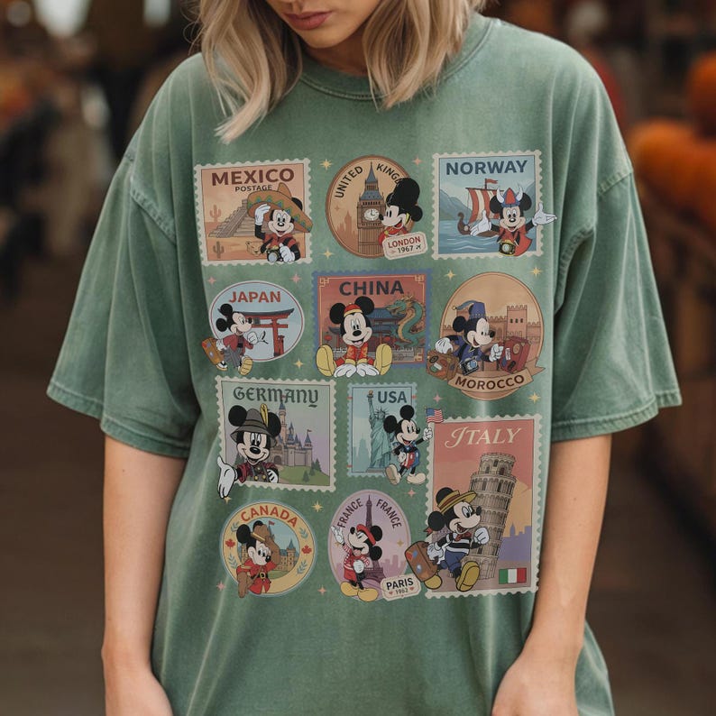 May include: Sage green t-shirt featuring Mickey Mouse postage stamp designs representing various countries, including Mexico, Japan, China, Germany, USA, Italy, and France. The stamps showcase Mickey in different outfits and settings.