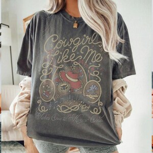 May include: A charcoal gray short-sleeve t-shirt featuring a graphic print with the text "Cowgirl like Me" and "Takes One to Know One". The design includes a red cowboy hat, rope borders, musical notes, and small floral accents in muted tones.