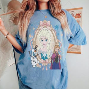 May include: Light blue t-shirt featuring a framed portrait of Elsa from Frozen, with Anna and Olaf. The design includes a pink bow, snowflakes, and floral accents. The shirt has short sleeves and a relaxed fit.