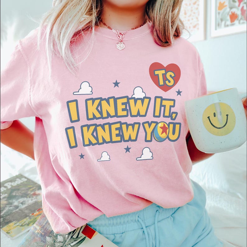 Op de afbeelding: Een lichtroze T-shirt met korte mouwen en de tekst I KNEW IT, I KNEW YOU in het geel met blauwe omlijning. Het ontwerp bevat een rood hart met de letters TS, kleine witte wolken, blauwe sterren en een kleurrijke balafbeelding met een rode ster.
