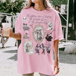 Pu&ograve; includere: T-shirt rosa chiaro con personaggi e testo di "Nightmare Before Christmas". Il design include Jack Skellington, Sally e altri personaggi iconici del film. Il testo recita "The Nightmare Before Christmas, The Pumpkin Queen & The Pumpkin King."