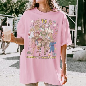 May include: A light pink t-shirt featuring a colorful graphic print of various animated characters in different outfits, including a space suit and cowgirl attire. The text TS and i KNEW iT. i KNEW YOU are printed in yellow on the front of the shirt.