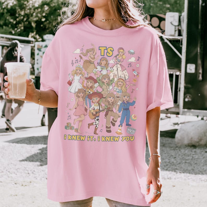 May include: A light pink t-shirt featuring a colorful graphic print of various animated characters in different outfits, including a space suit and cowgirl attire. The text TS and i KNEW iT. i KNEW YOU are printed in yellow on the front of the shirt.