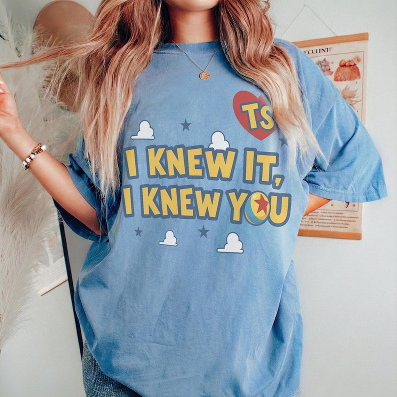 Op de afbeelding: Een lichtblauw t-shirt met korte mouwen en de gele tekst "I KNEW IT, I KNEW YOU", een rood hart met de letters "TS" en een kleurrijke balafbeelding. Het ontwerp bevat kleine witte wolkjes en donkergrijze sterren.
