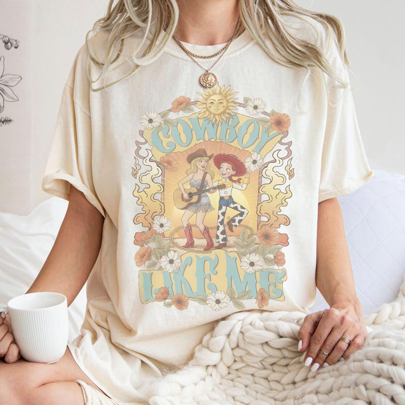 May include: A cream-colored t-shirt featuring a graphic print of two cartoon cowgirls, one playing a guitar, surrounded by flowers and a sun. The text COWBOY LIKE ME is printed in large blue letters. The shirt has short sleeves and a crew neckline.