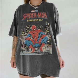 May include: Dark gray t-shirt featuring a graphic of Spider-Man in a crouching pose against a cityscape backdrop. The shirt has the text "MARVEL STUDIOS SPIDER-MAN BRAND NEW DAY" in red and white.