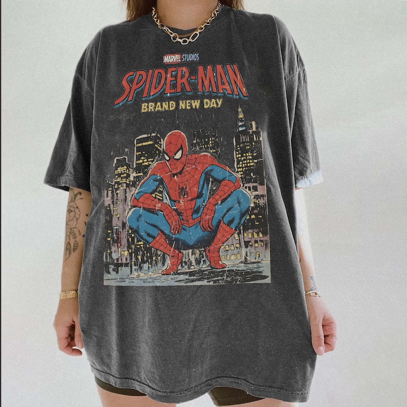 May include: Dark gray t-shirt featuring a graphic of Spider-Man in a crouching pose against a cityscape backdrop. The shirt has the text "MARVEL STUDIOS SPIDER-MAN BRAND NEW DAY" in red and white.
