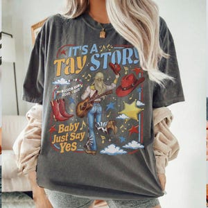 K&ouml;nnte beinhalten: Ein anthrazitfarbenes T-Shirt mit einem Grafikdruck mit dem Text IT'S A TAY STORY und Baby Just Say Yes. Das Design zeigt eine Person, die Akustikgitarre spielt, rote Cowboystiefel, einen roten Hut, einen gelben Stern, Wolken und Musiknoten.