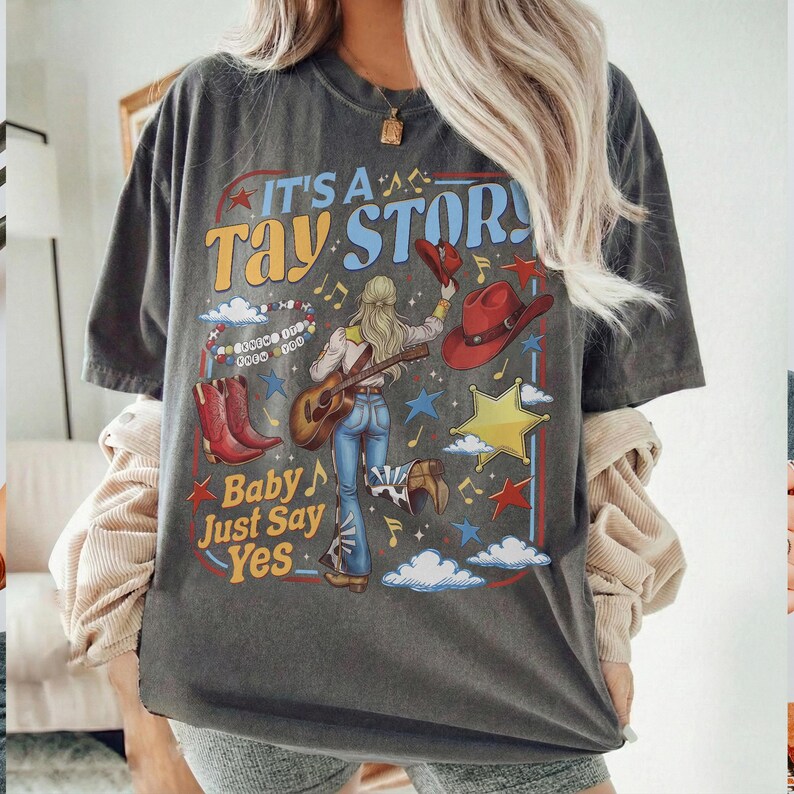 K&ouml;nnte beinhalten: Ein anthrazitfarbenes T-Shirt mit einem Grafikdruck mit dem Text IT'S A TAY STORY und Baby Just Say Yes. Das Design zeigt eine Person, die Akustikgitarre spielt, rote Cowboystiefel, einen roten Hut, einen gelben Stern, Wolken und Musiknoten.