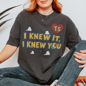 Op de afbeelding: Een antracietgrijs T-shirt met korte mouwen en de gele tekst I KNEW IT, I KNEW YOU. Het ontwerp bevat een rood hart met de letters TS, kleine witte wolkjes, blauwe sterren en een gele bal met een rode ster als afbeelding.