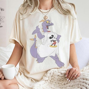 Peut inclure: T-shirt cr&egrave;me avec un dragon violet portant un t-shirt blanc avec un motif Mickey Mouse. Le dragon a des cornes et des griffes jaunes. Le t-shirt est de coupe ample.