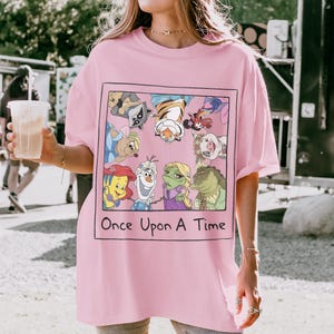 May include: Pink t-shirt featuring a framed graphic with cartoon characters and the text "Once Upon A Time." The graphic includes characters like Ariel, Olaf, and a raccoon. The shirt has short sleeves and a relaxed fit.