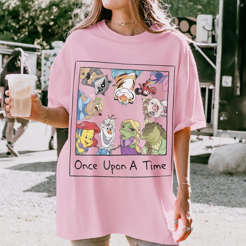May include: Pink t-shirt featuring a framed graphic with cartoon characters and the text "Once Upon A Time." The graphic includes characters like Ariel, Olaf, and a raccoon. The shirt has short sleeves and a relaxed fit.