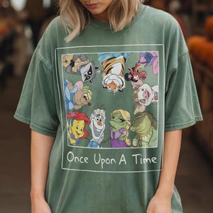 May include: Green t-shirt featuring a rectangular graphic with cartoon characters and the text "Once Upon A Time" in white. The graphic includes a raccoon, tiger, and other animated figures. The shirt has short sleeves and a relaxed fit.